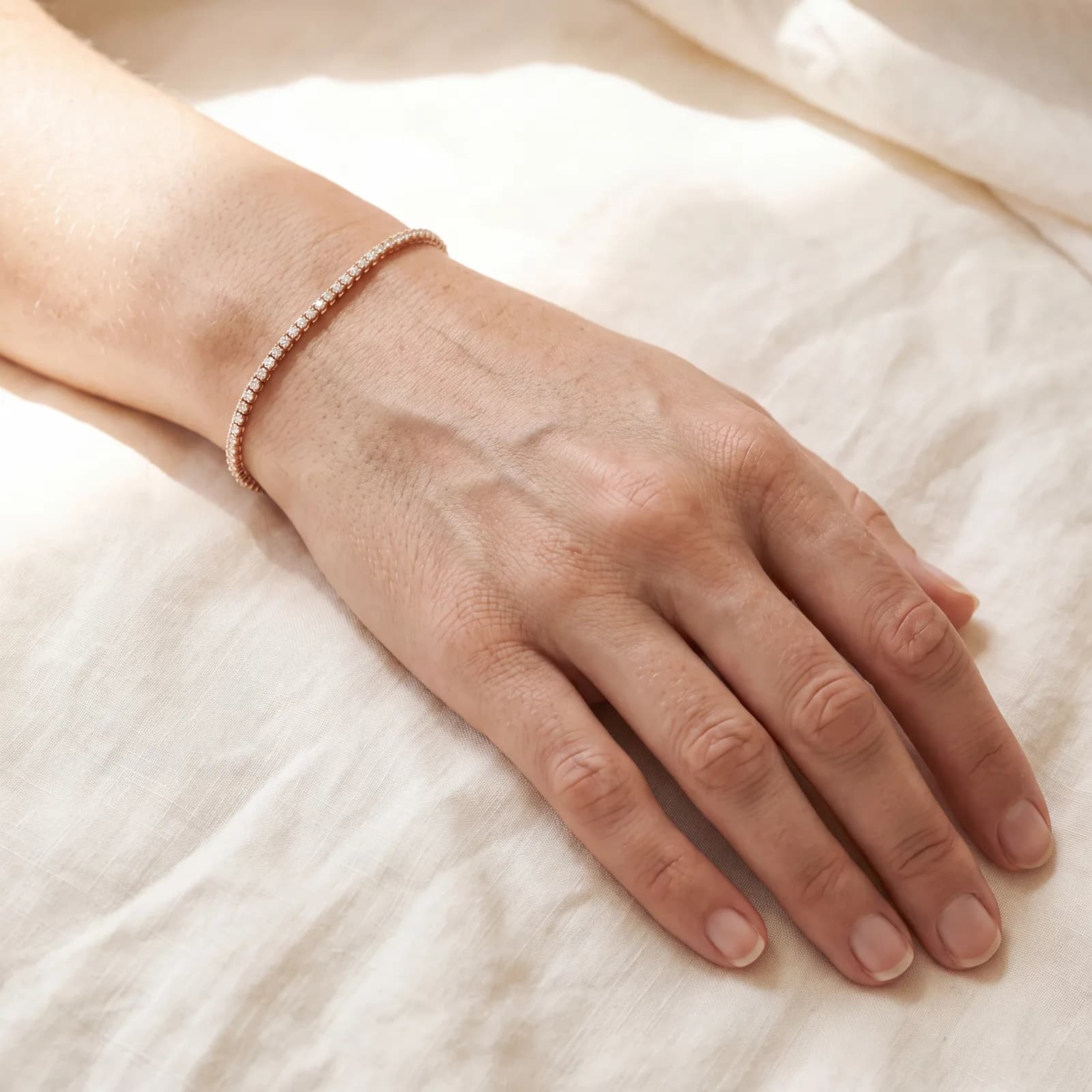 A close view of a wrist and hand resting on ivory linen, wearing a rose-gold-toned diamond line bracelet.
