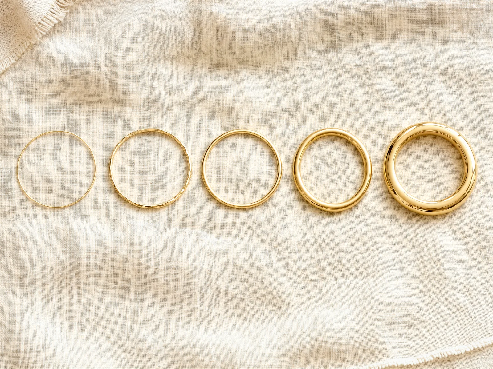 A stack of gold bangle bracelets in graduated gauges, from a thin 2mm bangle to a wide 10mm bangle, on a woven cream linen surface