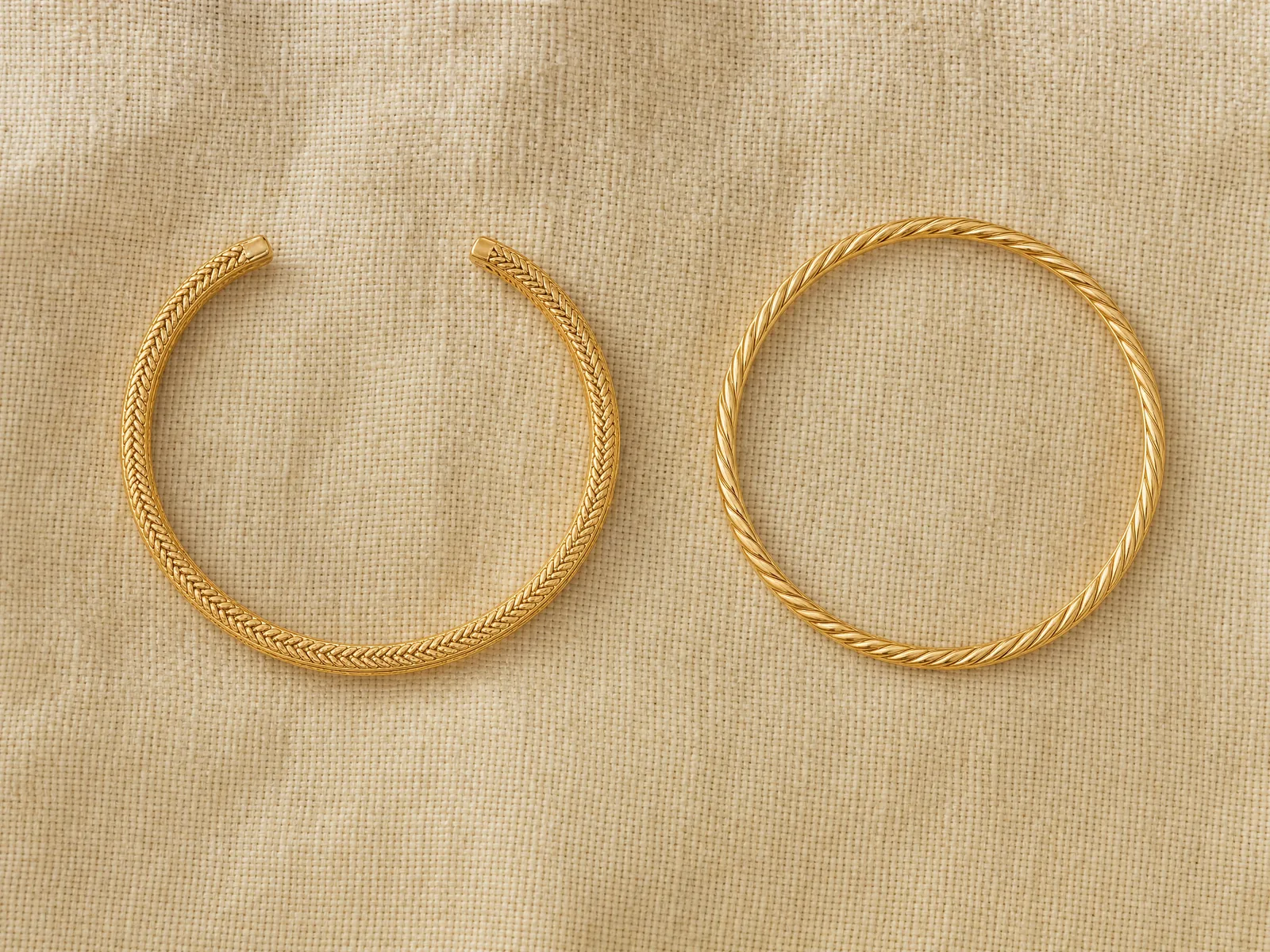 An open gold cuff bracelet standing on edge with its gap facing forward, beside a closed gold bangle standing on edge, on a woven cream linen surface