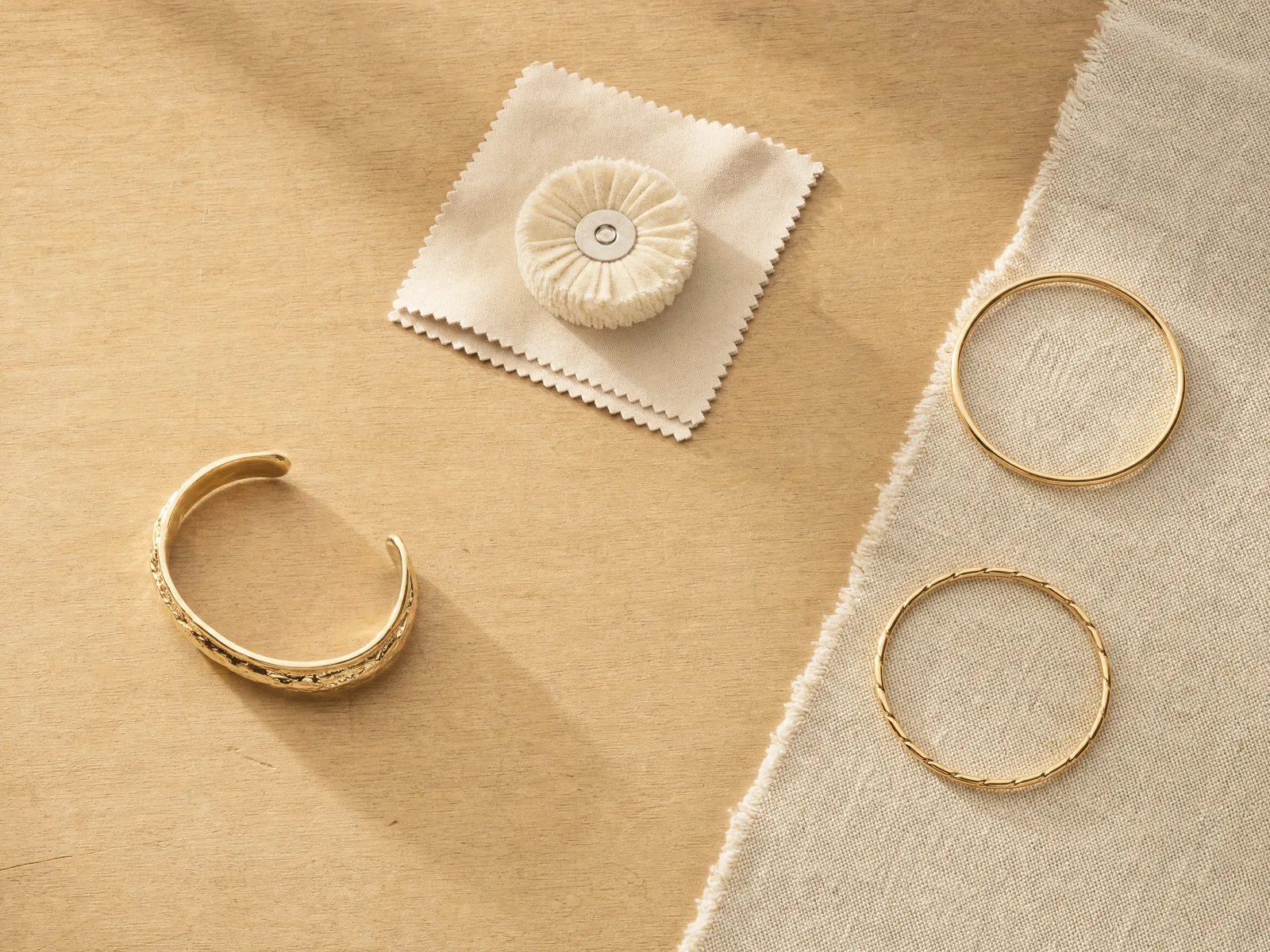 A gold cuff bracelet on a light wooden jeweler's workbench beside a polishing wheel and cloth, with finished gold bangles on woven cream linen alongside