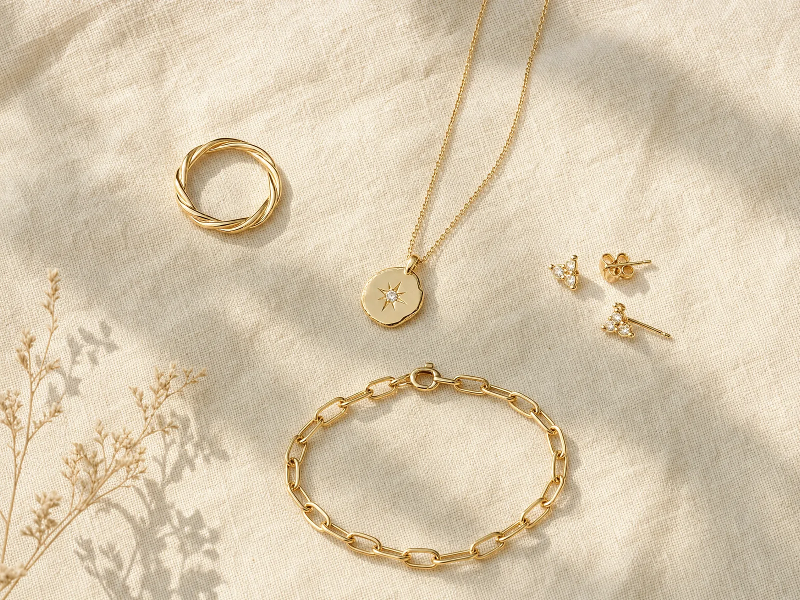 A flat lay of gold jewelry pieces — a ring, a bracelet, a pendant necklace and a pair of stud earrings — arranged on cream silk.