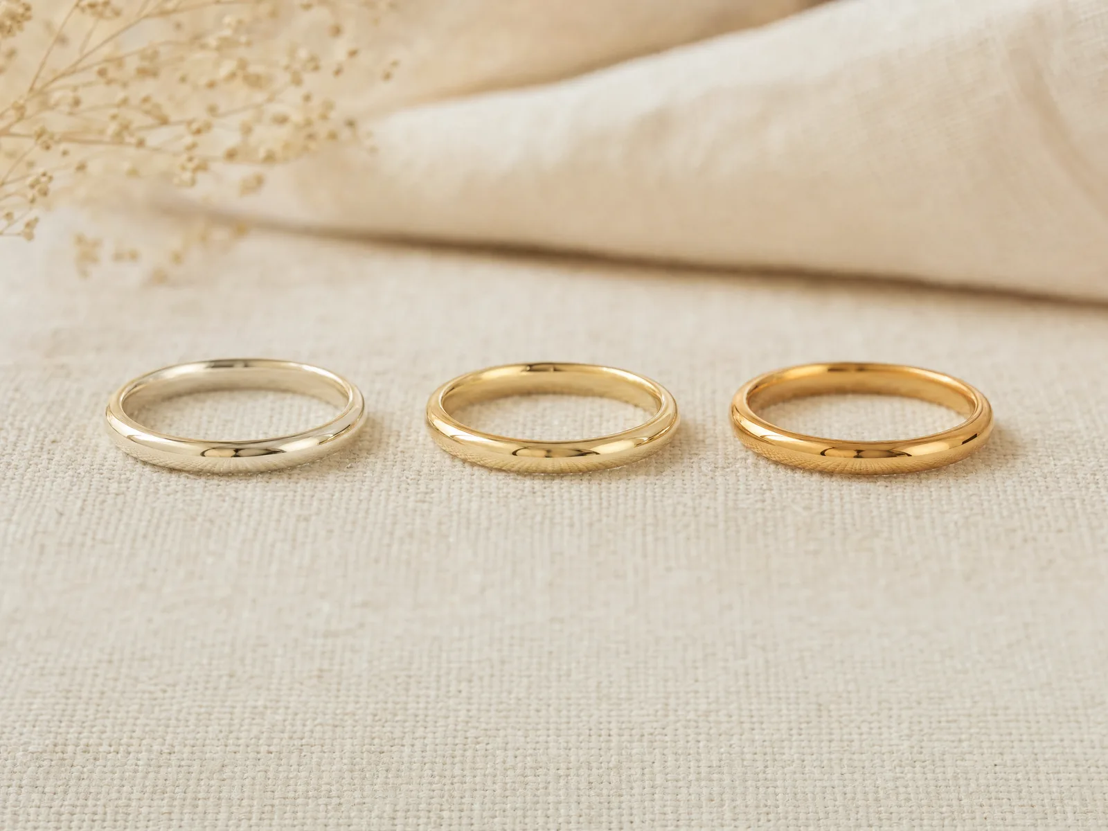 Three identical wedding band designs in 10K, 14K and 18K gold arranged in a row on woven cream linen in soft daylight.