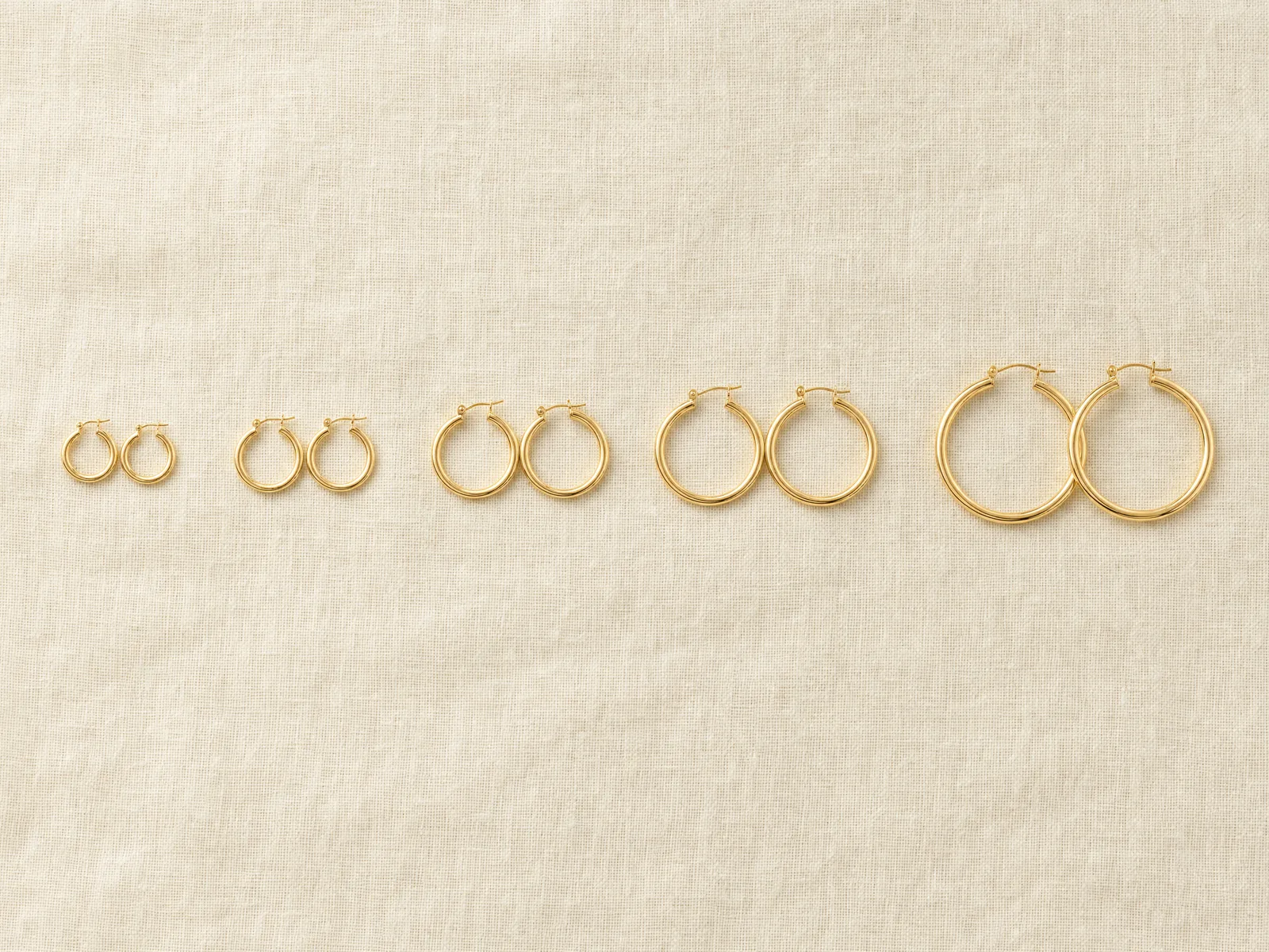 Five pairs of gold hoop earrings in ascending diameter — 10mm, 20mm, 30mm, 40mm and 50mm — laid out in a row on a woven cream linen surface in soft daylight.