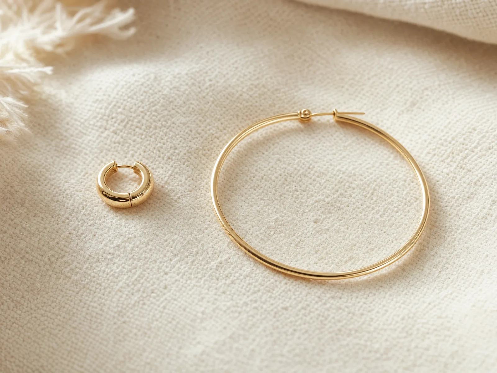 An 8mm gold huggie hoop and a 20mm gold hoop earring placed side by side on a woven cream linen surface, showing the scale difference between daily-wear and statement sizes.