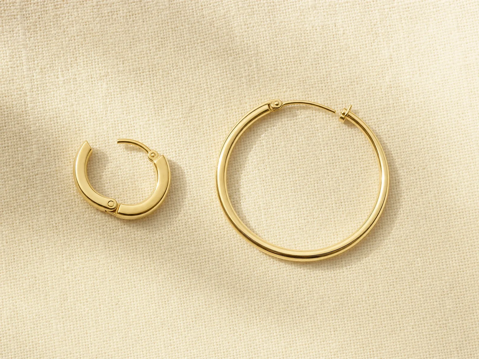 A small hinged huggie hoop earring, open at its clasp, next to a larger 30mm click-top hoop earring, on a woven cream linen surface in soft daylight.