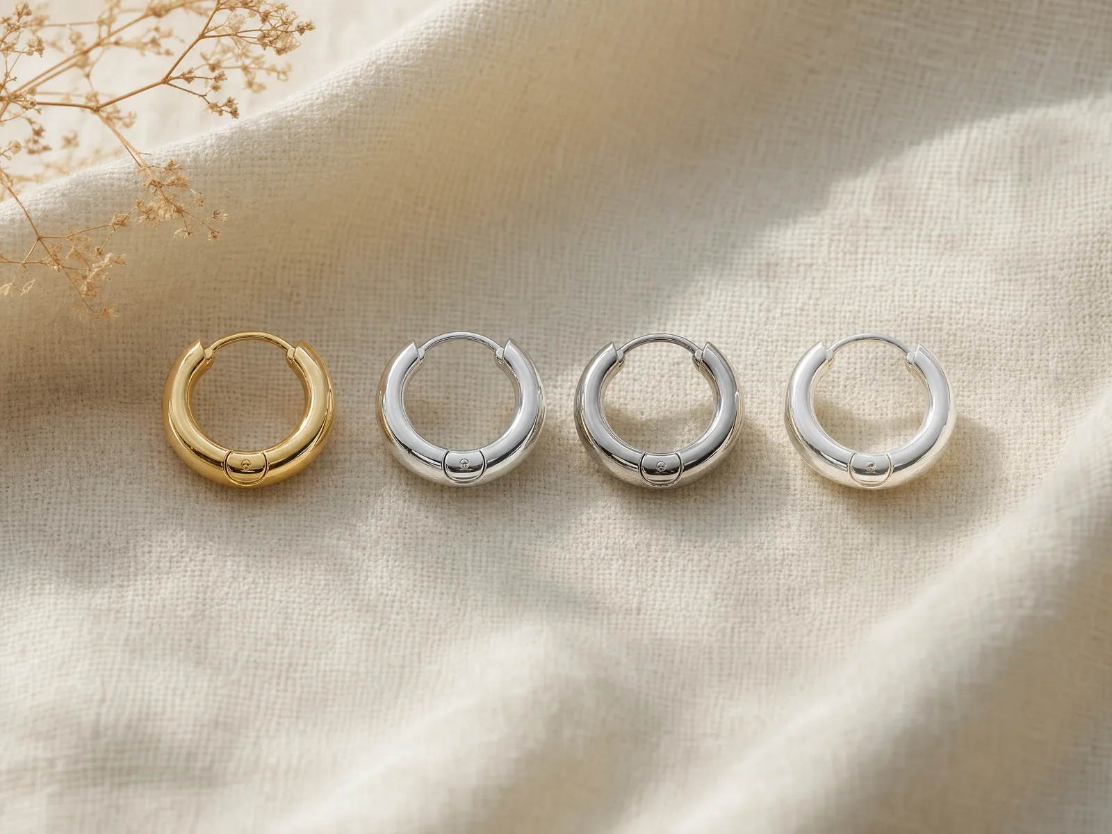 Four hoop earrings in different metals — 14K yellow gold, 14K white gold, platinum, and sterling silver — arranged in a row on a woven cream linen surface in soft daylight.