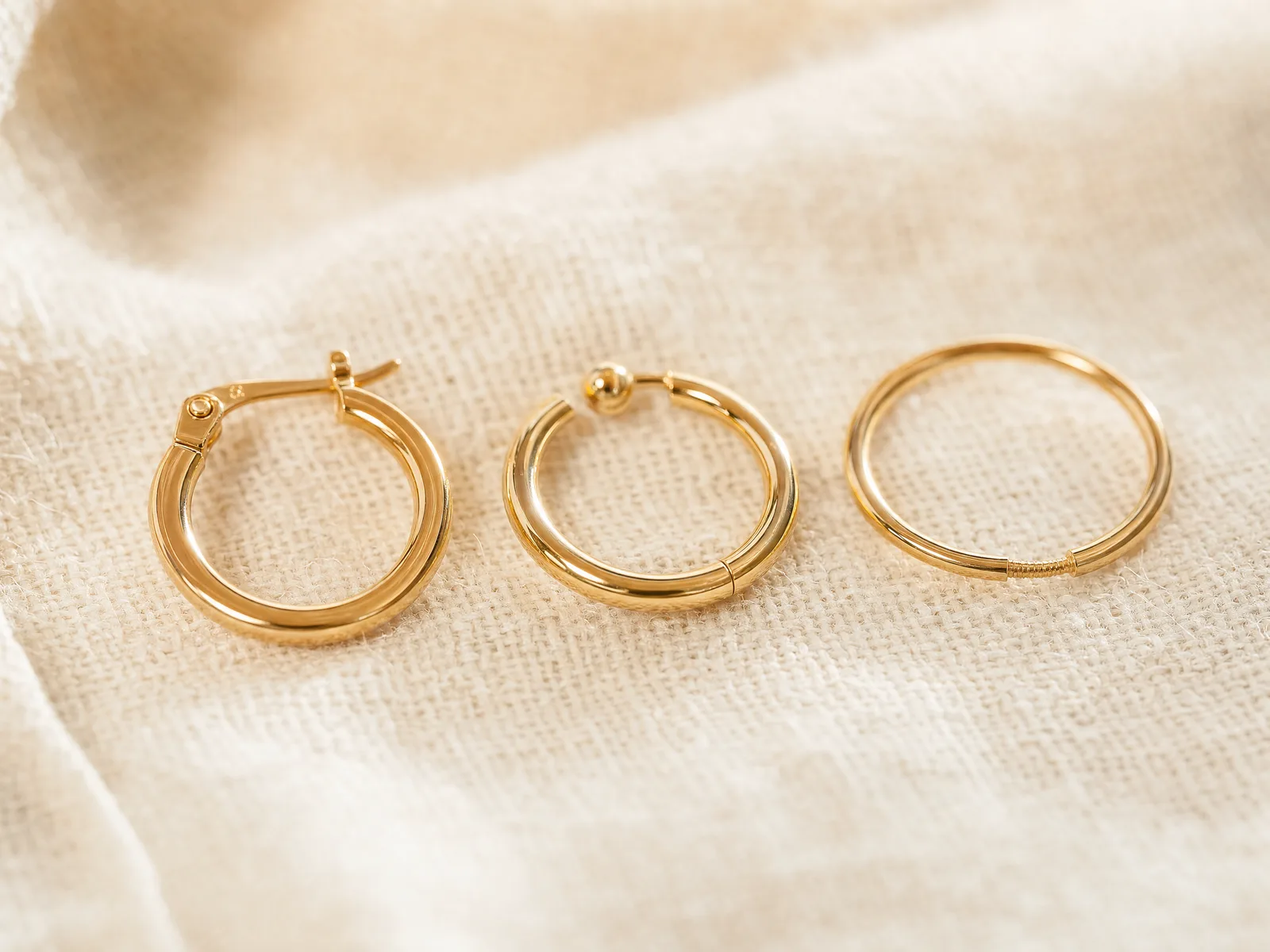 Three hoop earring closures side by side on a woven cream linen surface in soft daylight — a hinged clasp, a click-top ball closure, and an endless threaded wire.