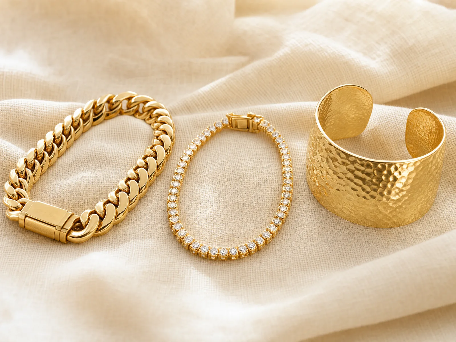 Three men's gold bracelets side by side on a woven cream linen surface — a Cuban link bracelet, a diamond tennis bracelet, and a wide hammered cuff