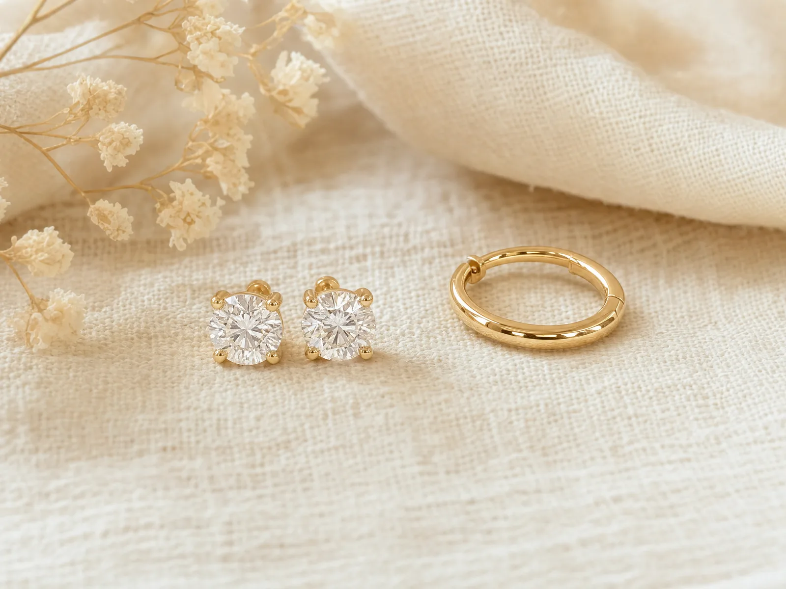 A pair of men's round brilliant diamond stud earrings in 4-prong settings beside a single gold hoop earring on a woven cream linen surface
