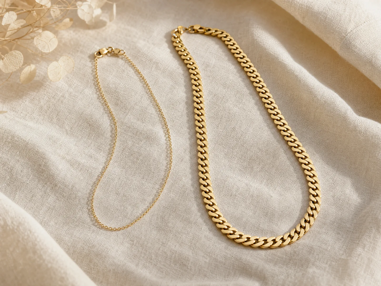 An 18-inch women's cable chain and a 22-inch men's Cuban link chain laid side by side on a woven cream linen surface in soft daylight, showing the length and gauge difference.
