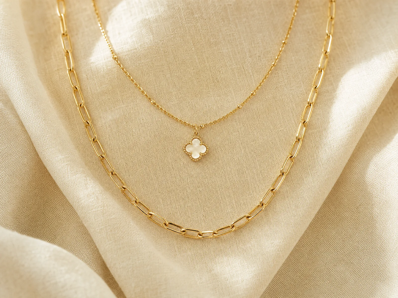 Three necklaces layered together at 16, 18 and 20-inch lengths — a delicate cable chain, a pendant chain, and a Cuban link chain — arranged on a woven cream linen surface in soft daylight.