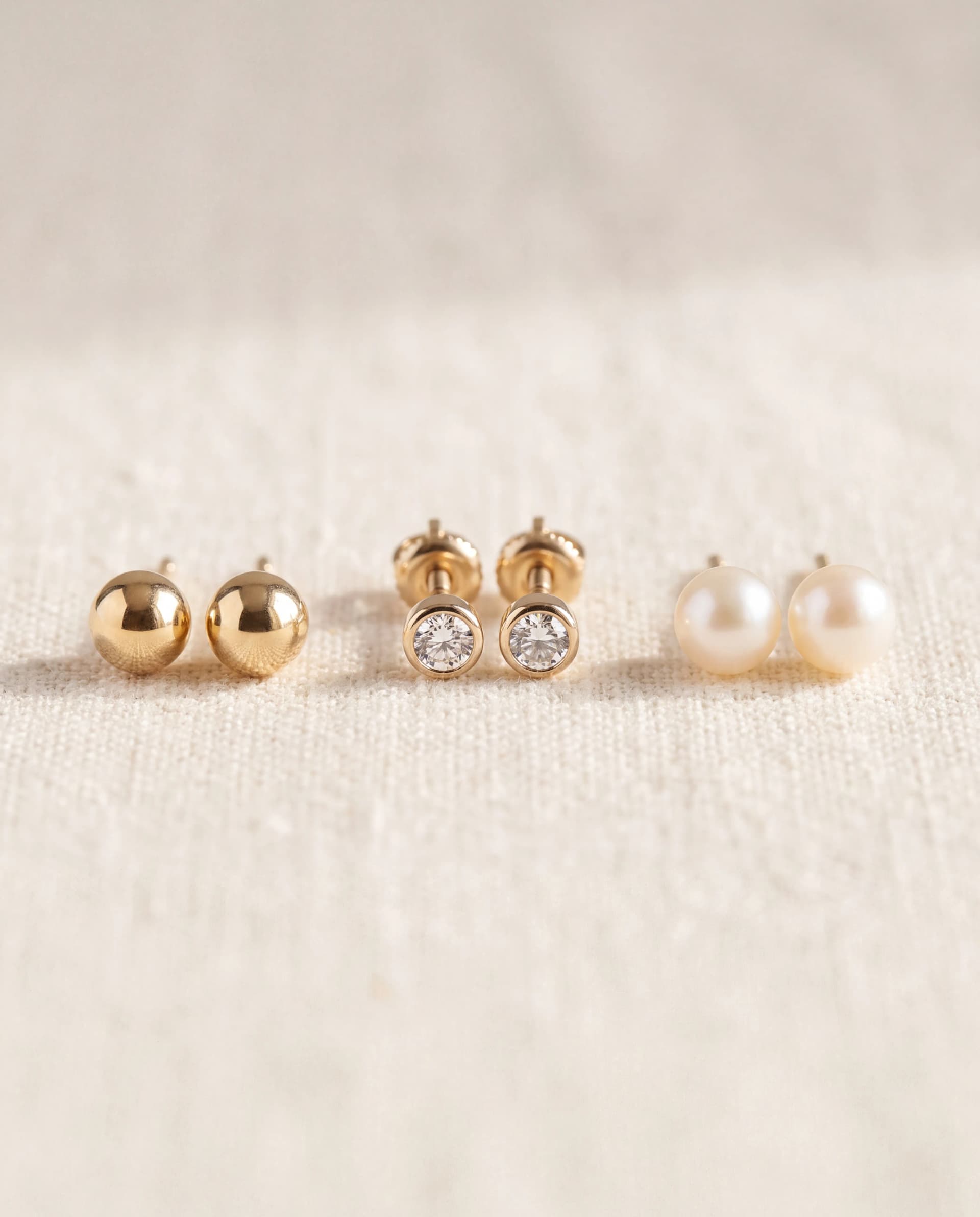 Three pairs of stud earrings on linen: a gold ball stud, a bezel-set diamond stud and a pearl stud