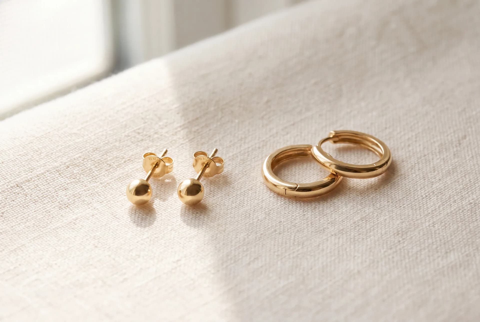 A pair of plain gold starter studs beside a pair of small polished gold huggie hoops on ivory linen
