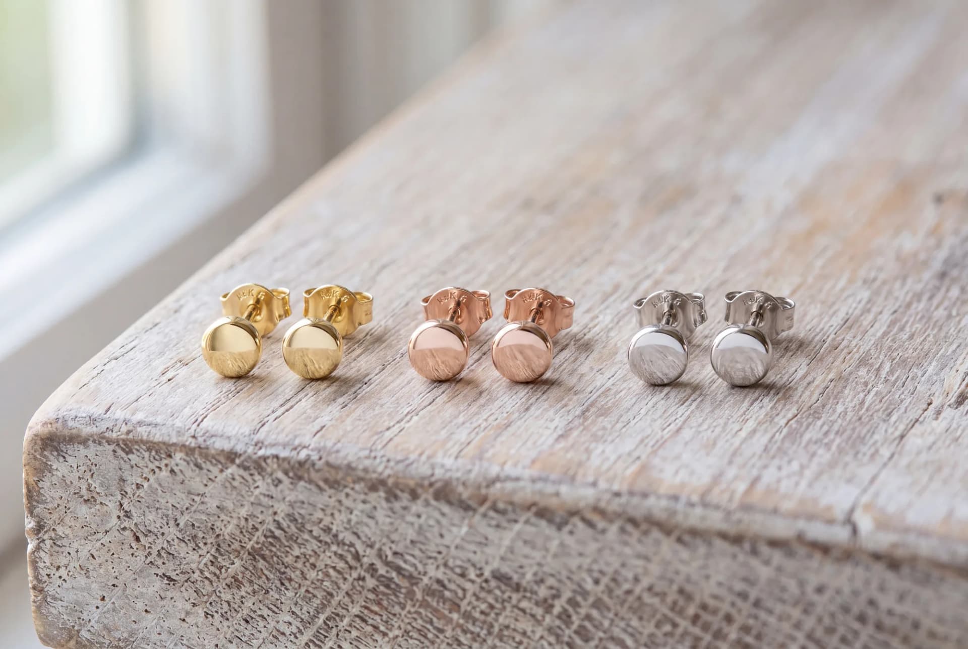 Three pairs of plain polished round stud earrings in yellow, rose and white gold on a pale wood surface