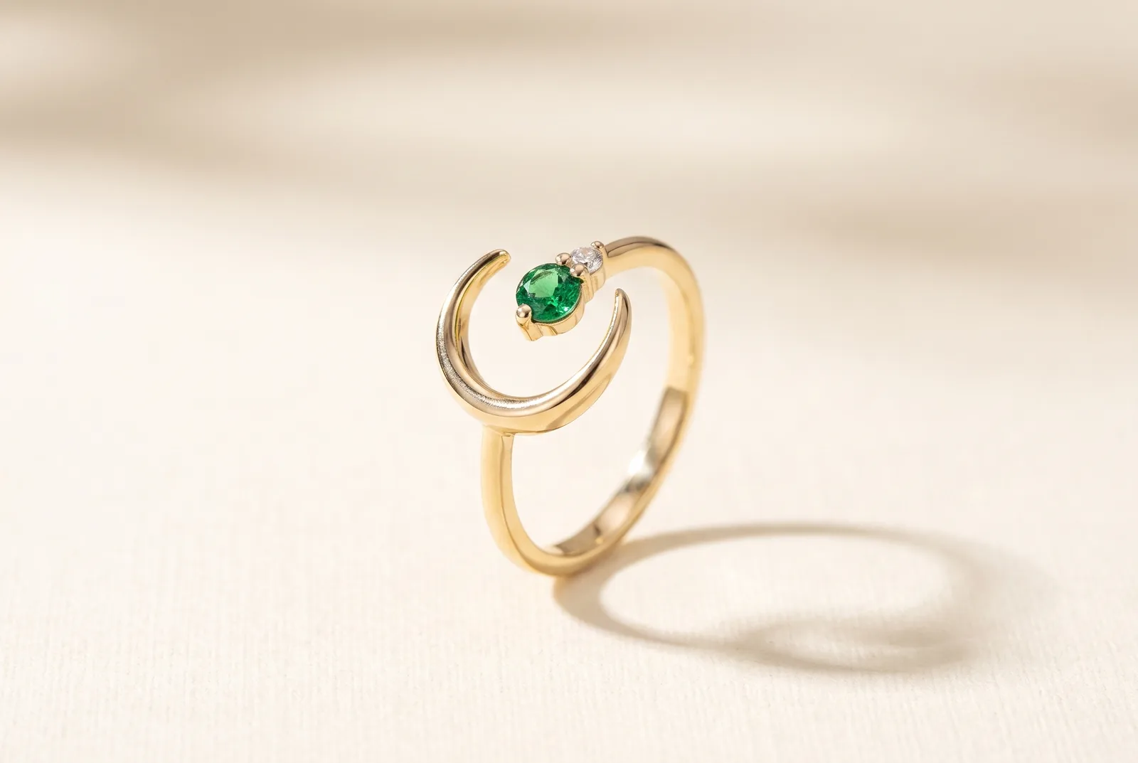 A design study for a yellow gold ring on a cream surface: an open crescent-moon form cradling a round emerald-green stone with a small round diamond at its shoulder, on a plain polished band