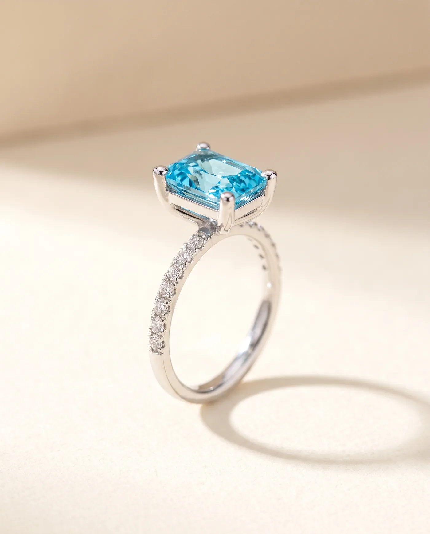 A design study for a white metal ring on a warm cream surface: a vivid sky-blue emerald-cut stone held in four prongs above an open basket, on a slim band lined with a single row of pave diamonds