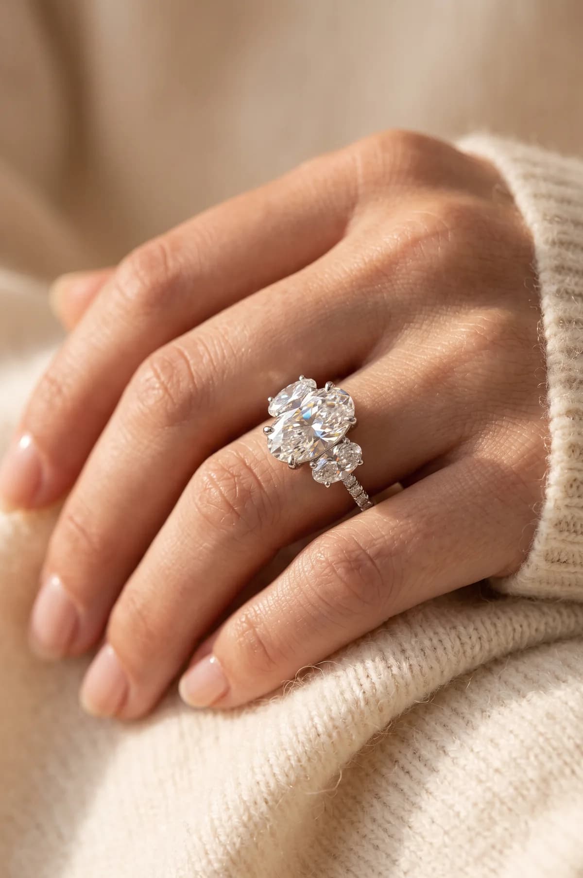 Close-up of clasped hands against cream knit, wearing a platinum three-stone engagement ring with an oval diamond centre and oval side stones