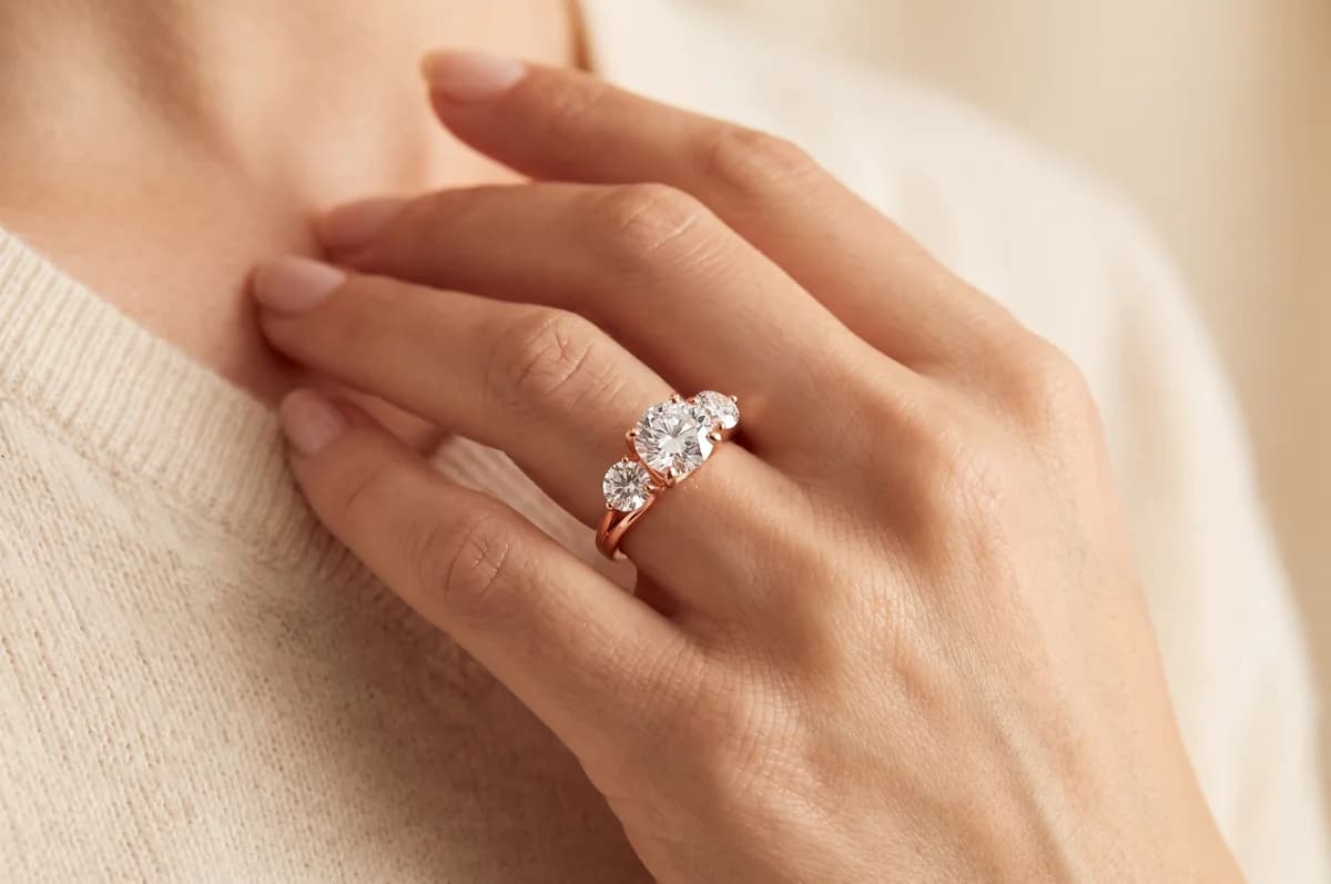 Close-up of a hand resting at the collar of a cream sweater, wearing a rose gold three-stone ring with a round centre diamond