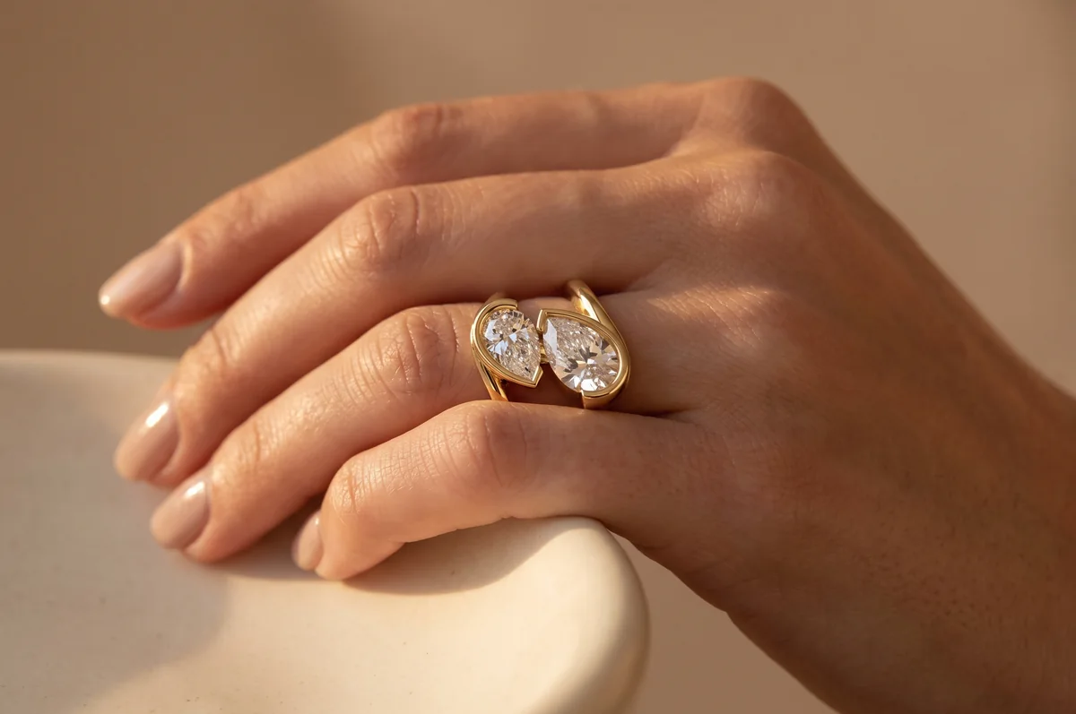 Close-up of a hand resting on a ceramic surface, wearing a yellow gold two-stone ring with two bezel-set pear-shaped diamonds