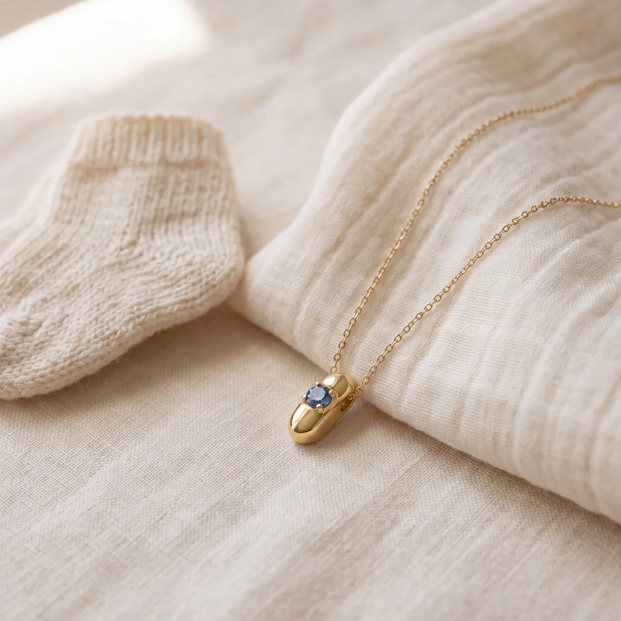 A gold birthstone pendant necklace laid beside a knit baby sock on linen.