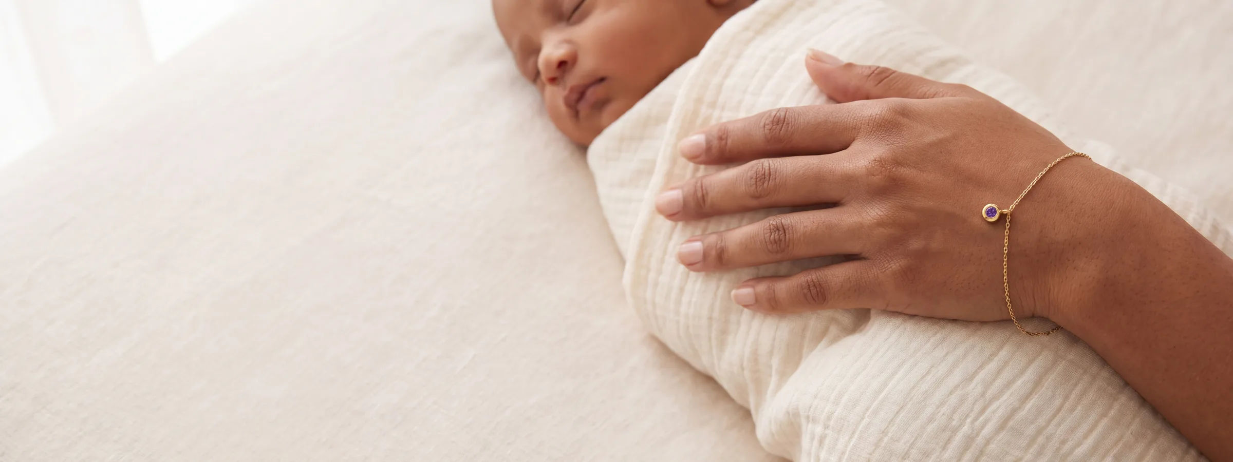 A mother's hand resting on her swaddled newborn, wearing a gold birthstone bracelet.