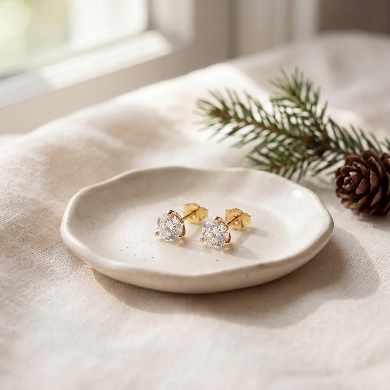 A pair of diamond stud earrings in a small ceramic dish on cream linen, beside a pine sprig and a pinecone