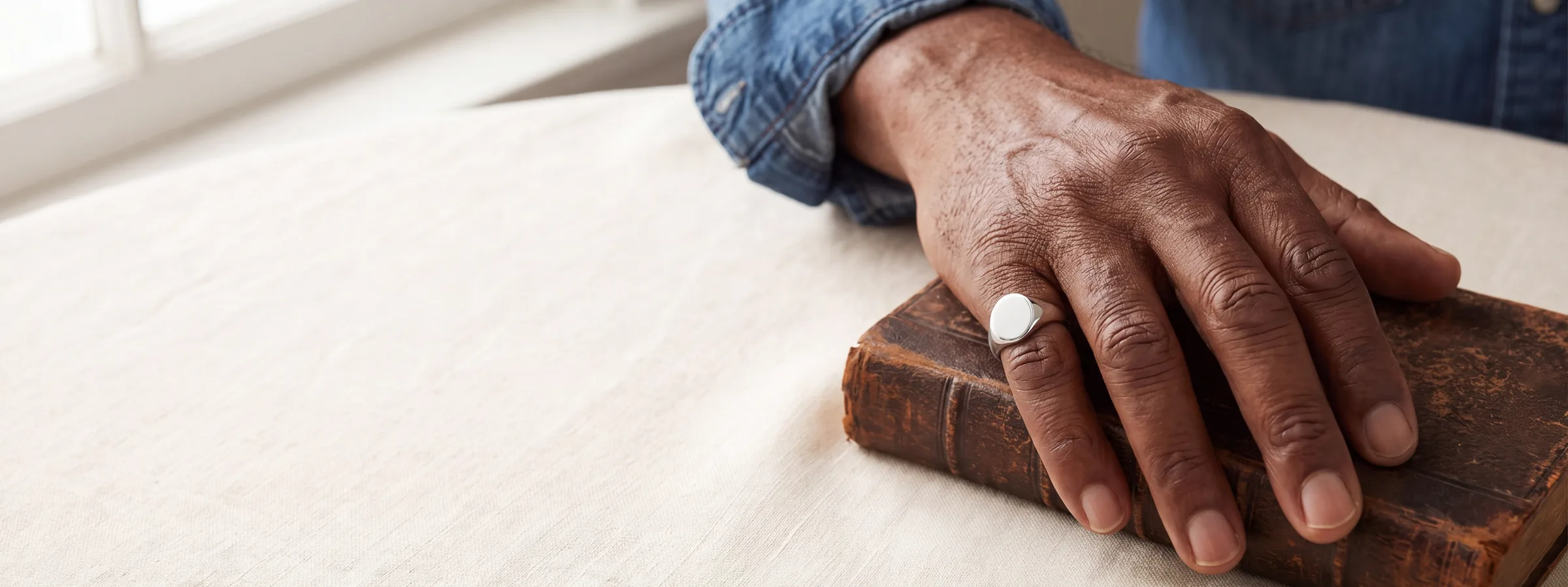 A hand resting on a worn leather book, wearing a plain oval signet ring