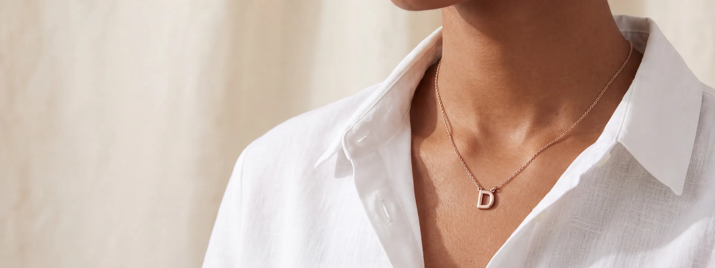 A rose gold initial pendant on a fine chain, worn open at the collar of a white shirt