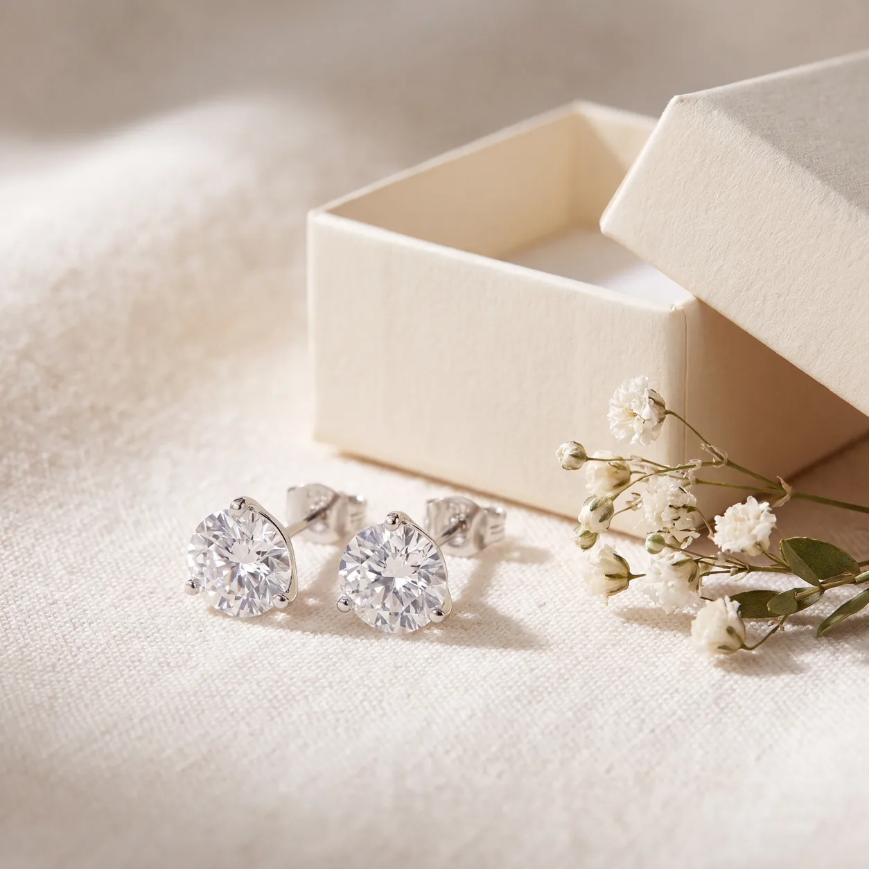 A pair of diamond stud earrings on linen beside a small open box and sprigs of baby's breath.