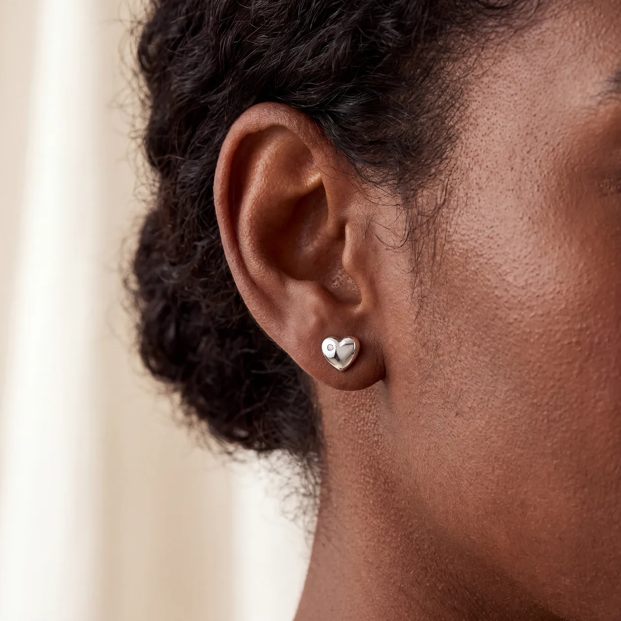A close-up of a woman's ear wearing a small silver heart stud earring with a diamond accent