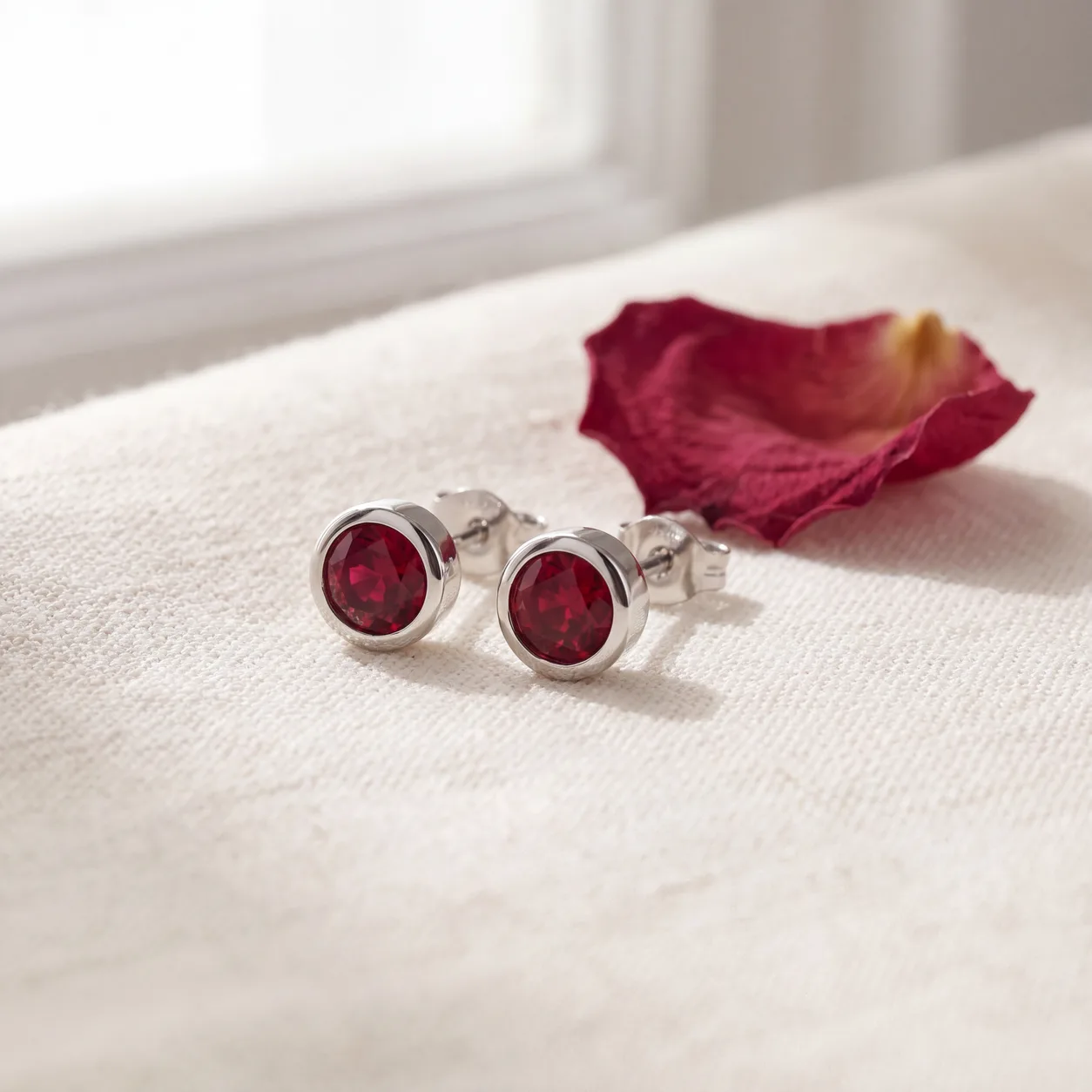 A pair of ruby stud earrings resting on cream linen fabric beside a dried rose petal