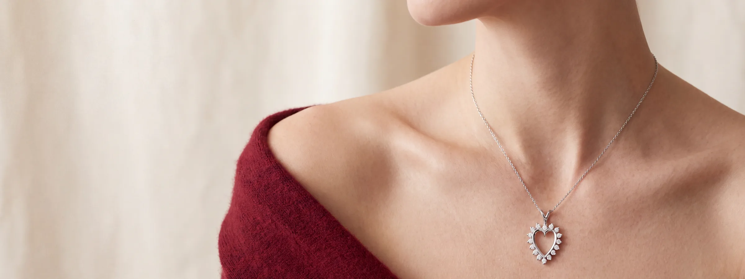 A woman's shoulder and collarbone wearing a silver open-heart pendant necklace, a red sweater draped off one shoulder