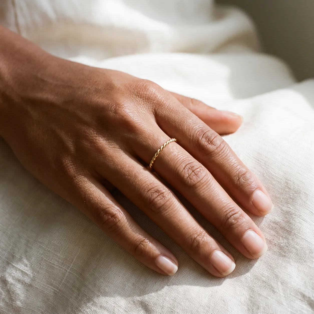 A woman's hand wearing one thin twisted gold ring band, resting on cream linen