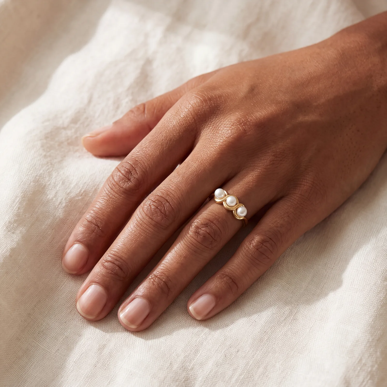 A hand resting on cream linen, wearing a yellow gold ring set with three round white pearls