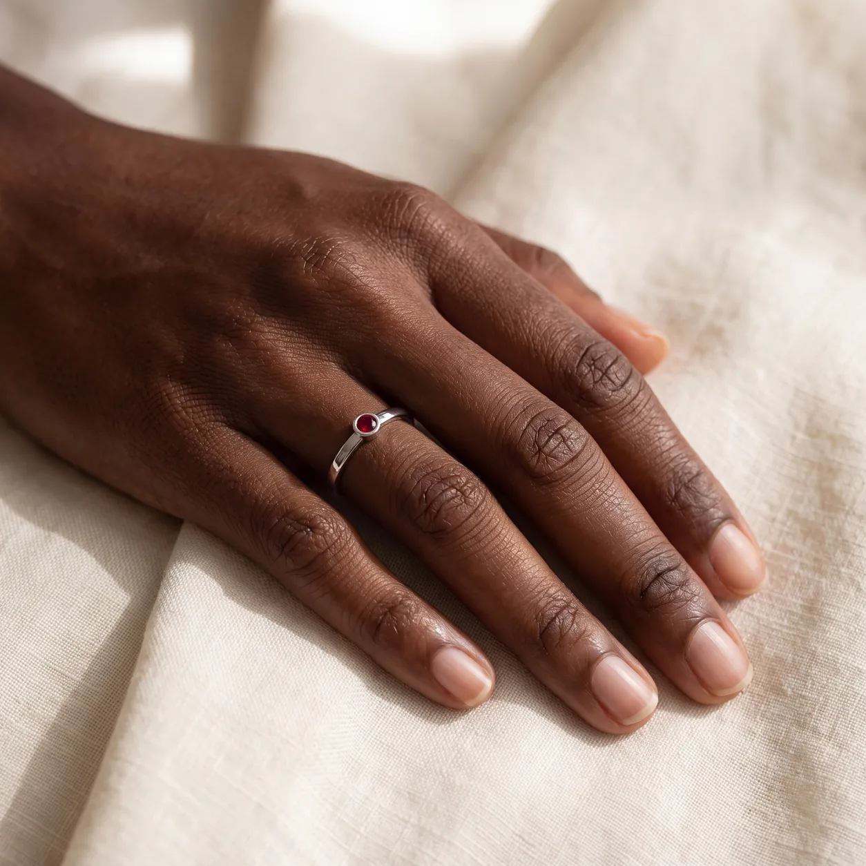 A hand wearing a thin ring set with a small round red stone, resting on linen