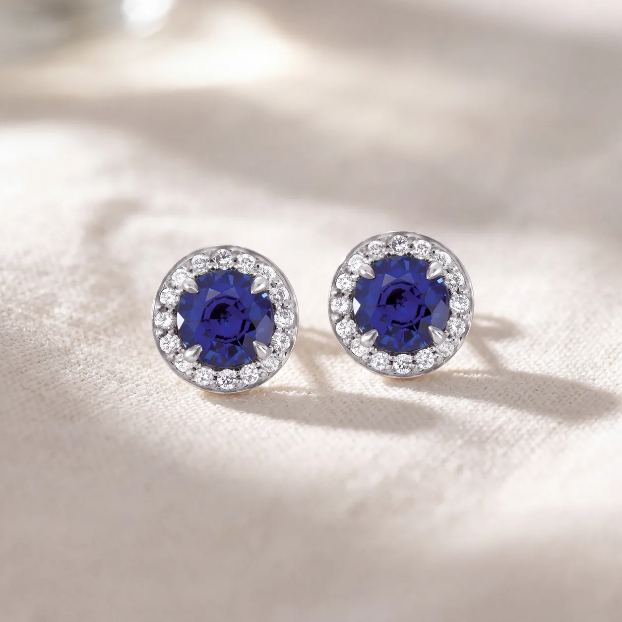 A pair of stud earrings, each a blue stone framed by a halo of diamonds, resting on linen