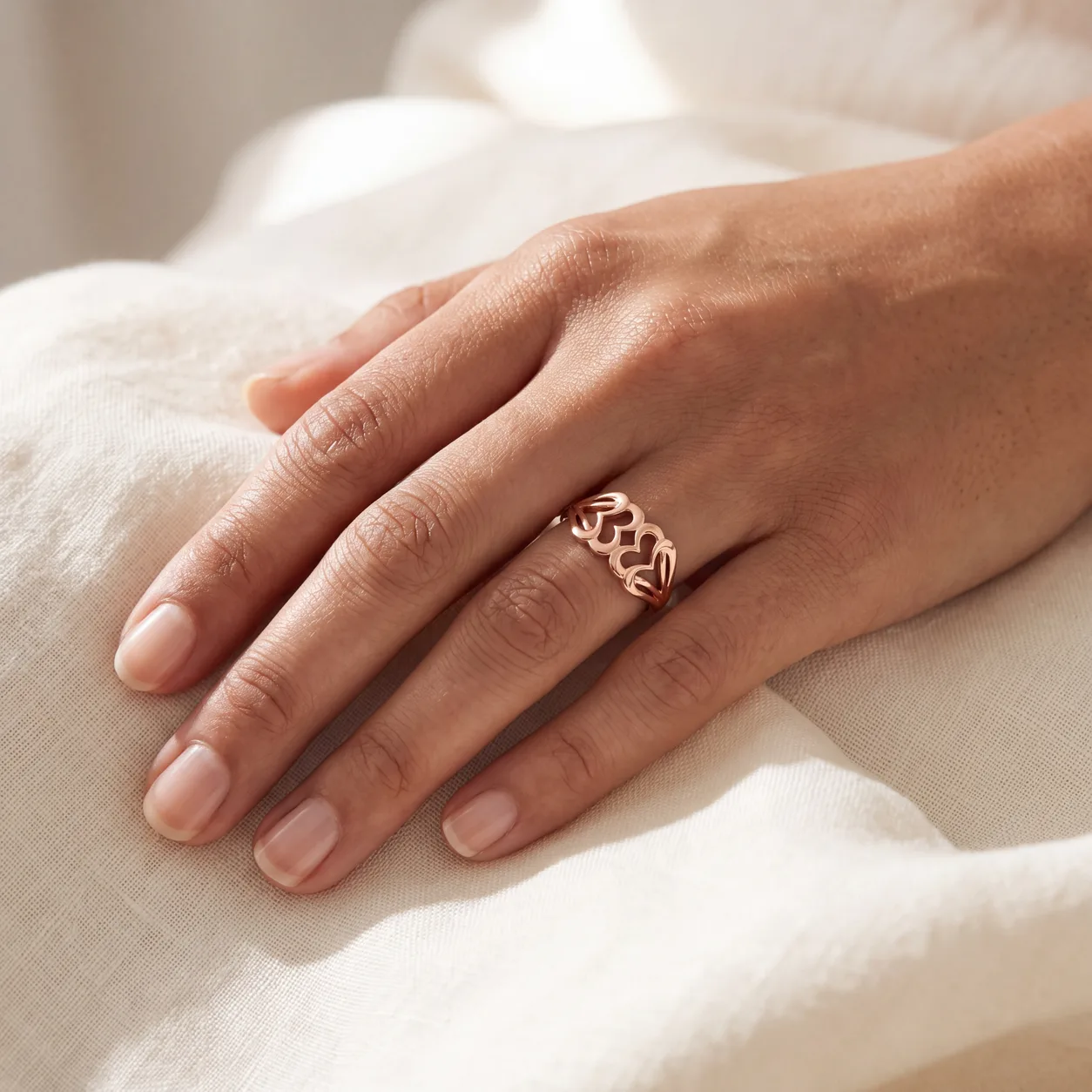 A woman's hand resting on cream linen wearing a rose gold ring set with three open heart shapes