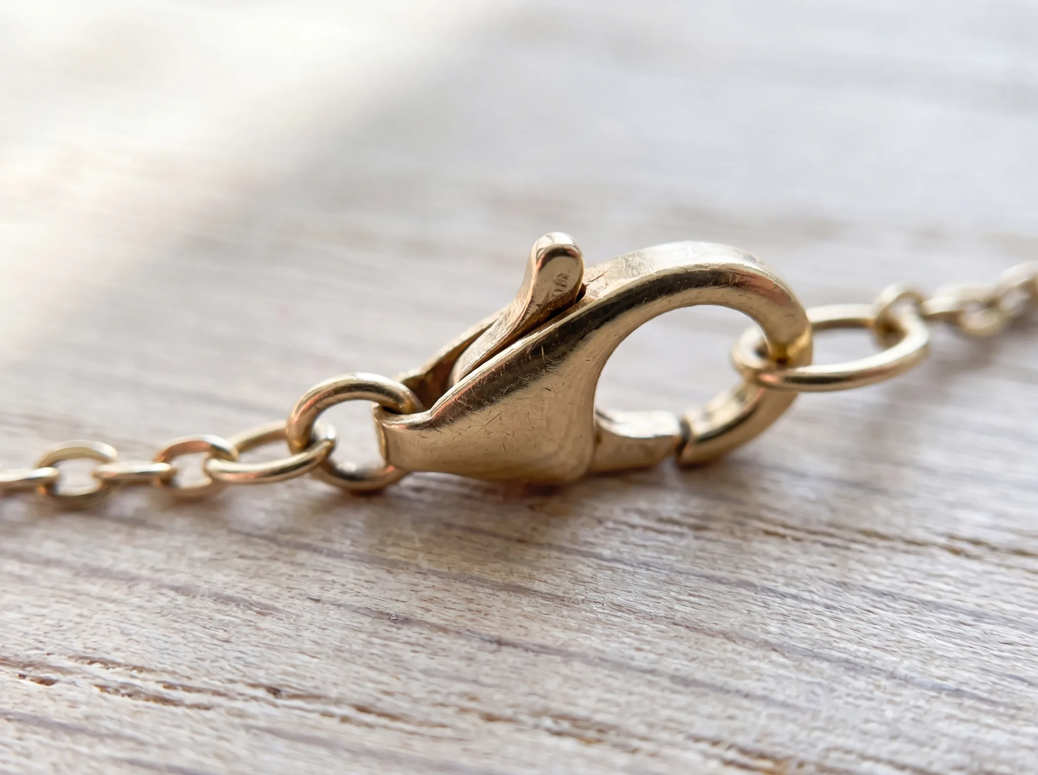 A worn gold lobster clasp with its spring lever sprung slightly open, resting on a pale weathered oak surface