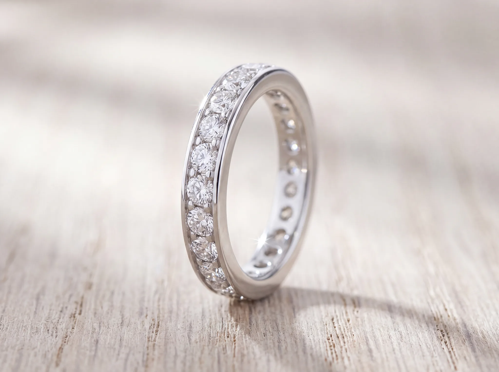 A white gold eternity band set with round diamonds continuously around its full circumference, standing on a pale wood surface