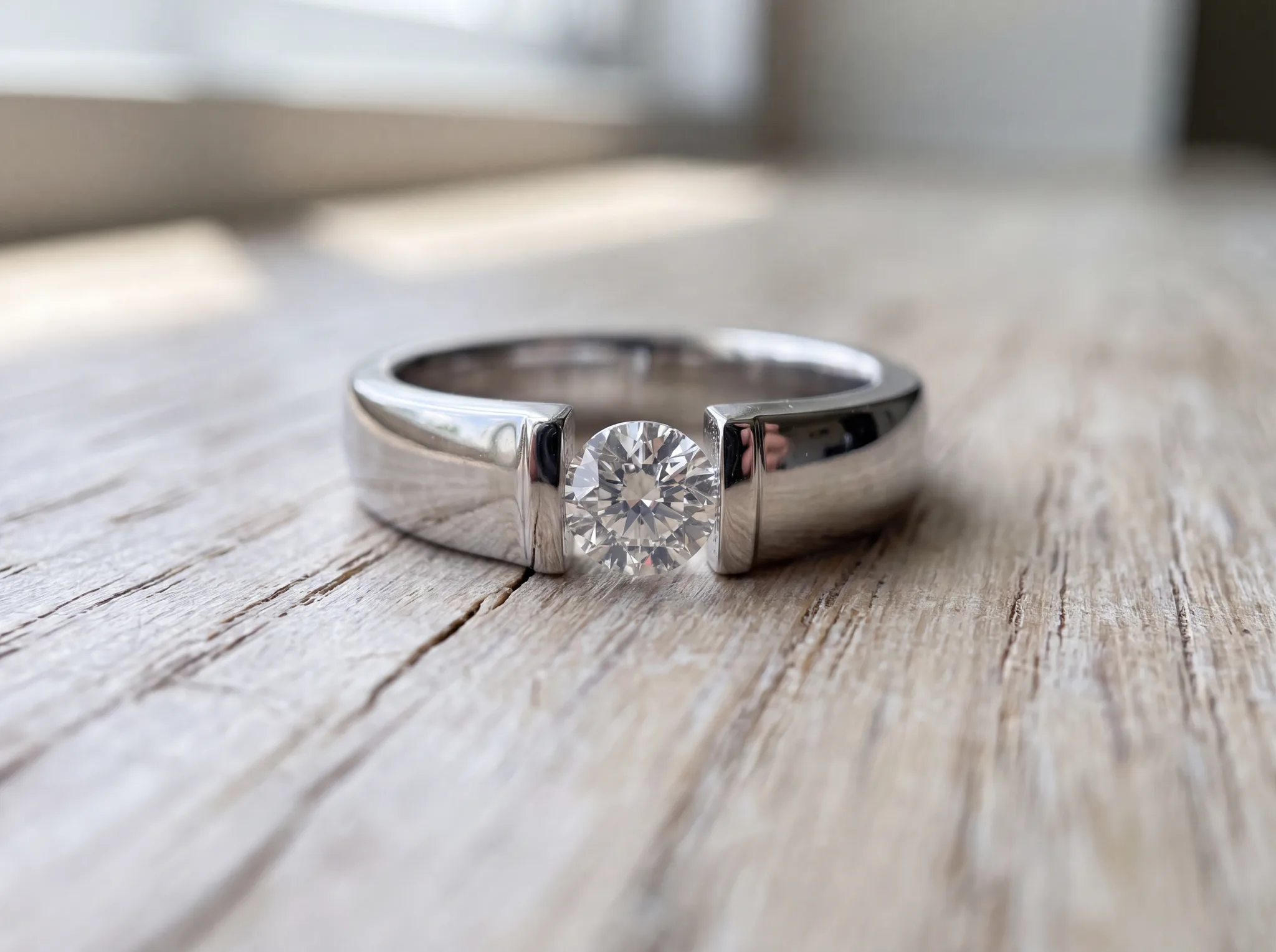 A tension-set engagement ring with a round diamond suspended between the polished ends of an open white gold band, on a pale wood surface