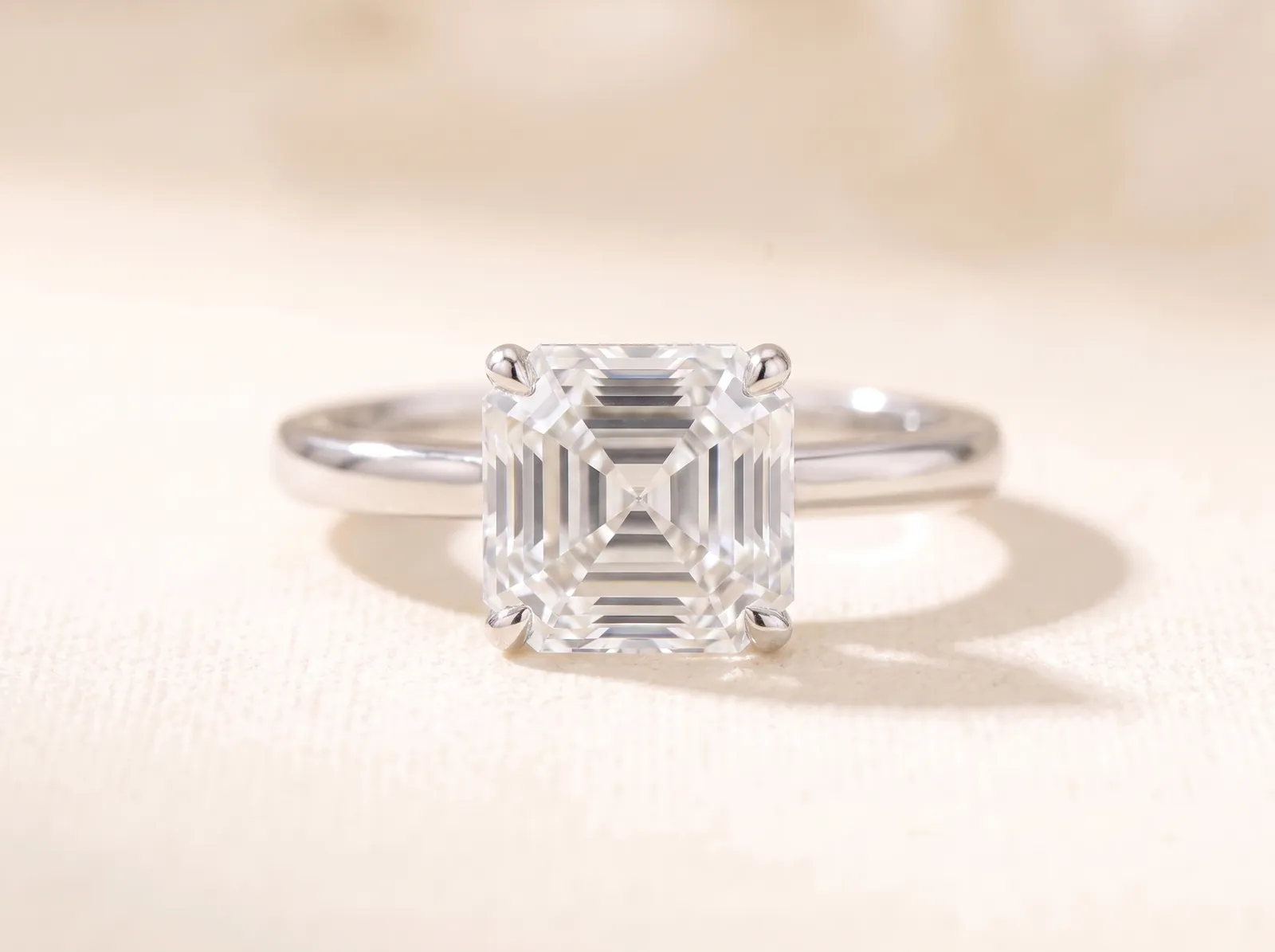The same platinum solitaire lying on the cream surface, now holding an asscher-cut diamond, a cropped-corner square with concentric step facets, facing the camera.