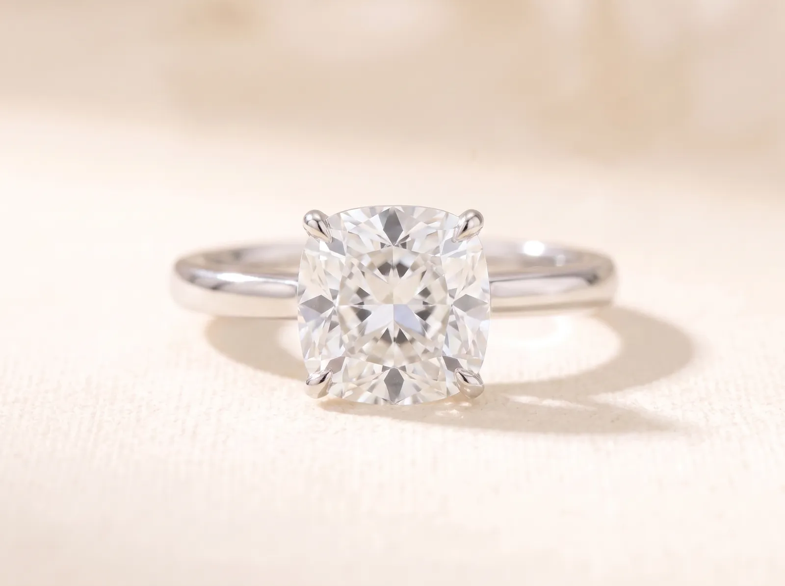 The same platinum solitaire lying on the cream surface, now holding a cushion-cut diamond, its softly rounded square outline facing the camera.