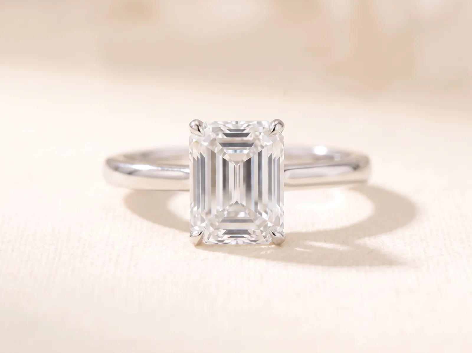 The same platinum solitaire lying on the cream surface, now holding a rectangular emerald-cut diamond with cropped corners and long parallel step facets facing the camera.