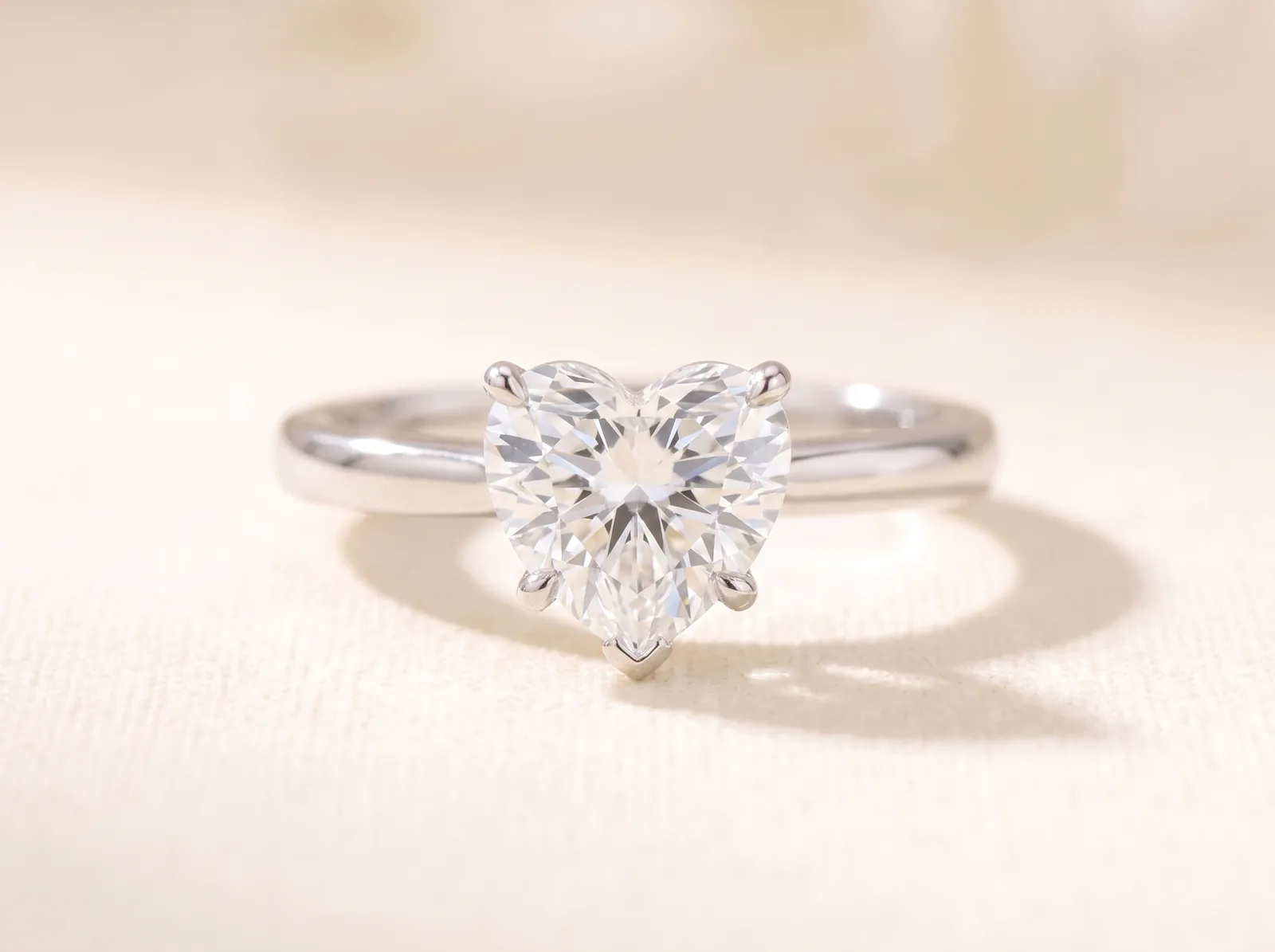 The same platinum solitaire lying on the cream surface, now holding a heart-shaped diamond with a defined cleft above and a point below, facing the camera.
