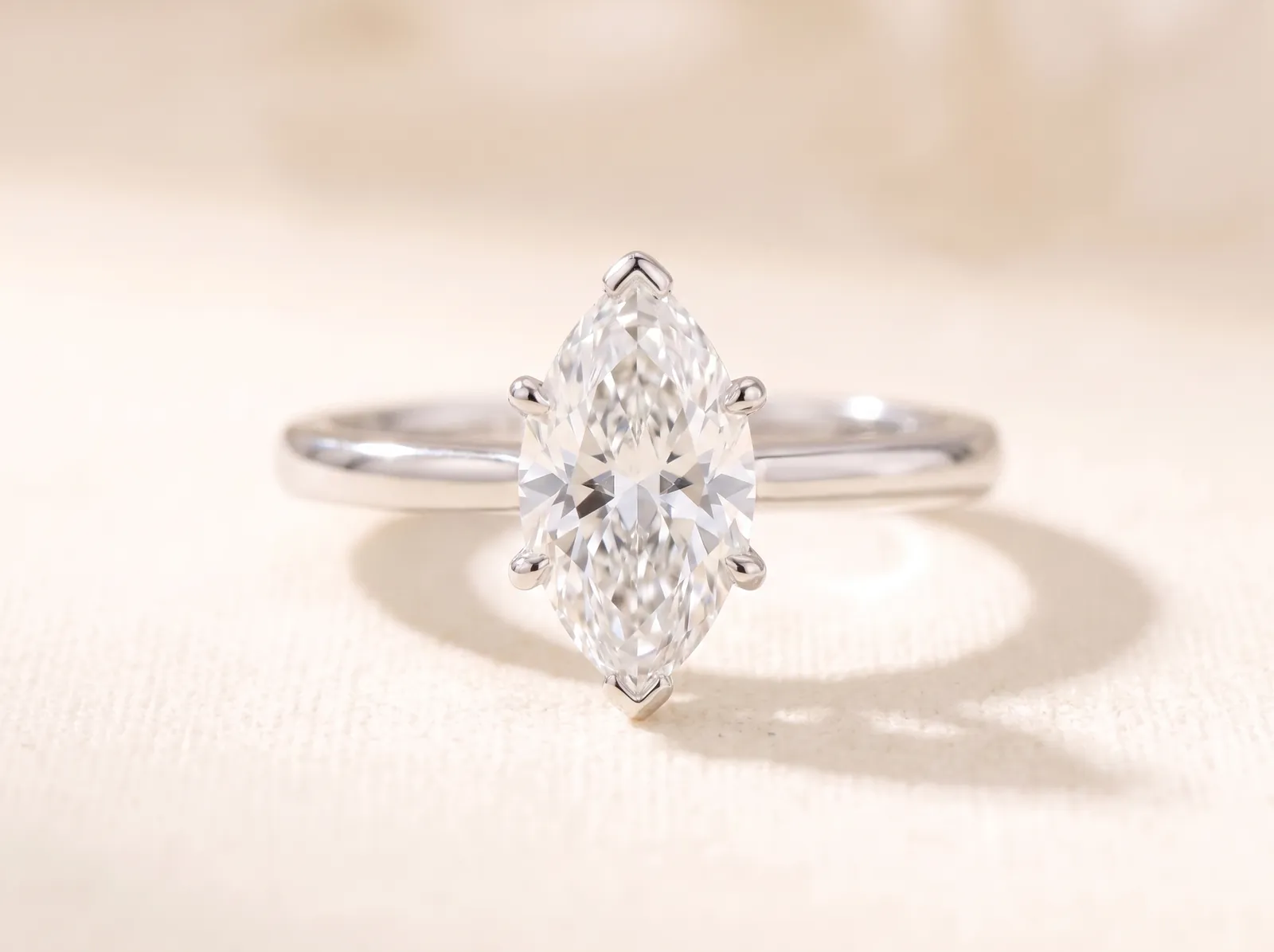 The same platinum solitaire lying on the cream surface, now holding a marquise diamond, long and narrow with a point at both ends, facing the camera.