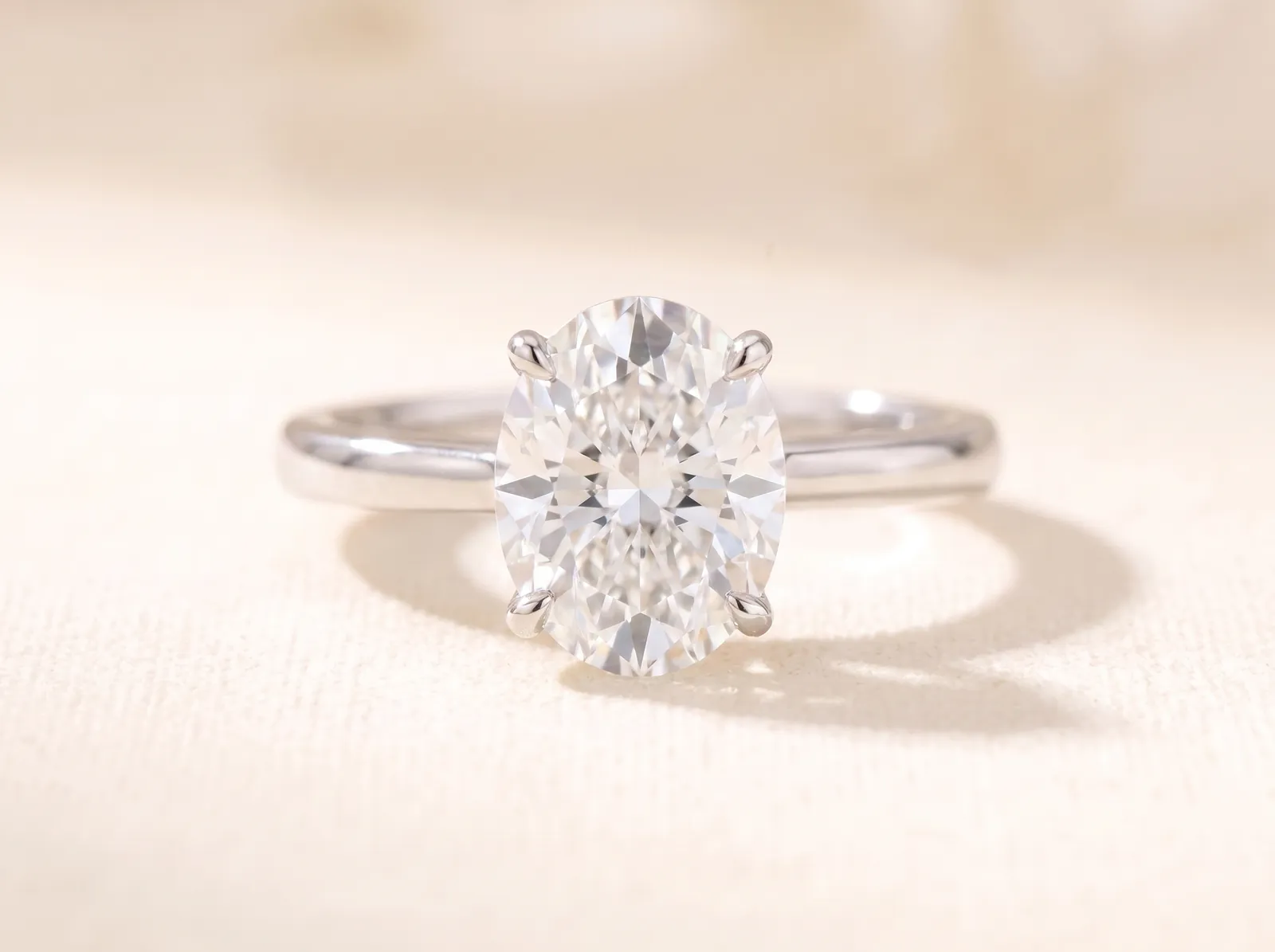 The same platinum solitaire lying on the cream surface, now holding an elongated oval diamond, its smooth ellipse outline facing the camera.