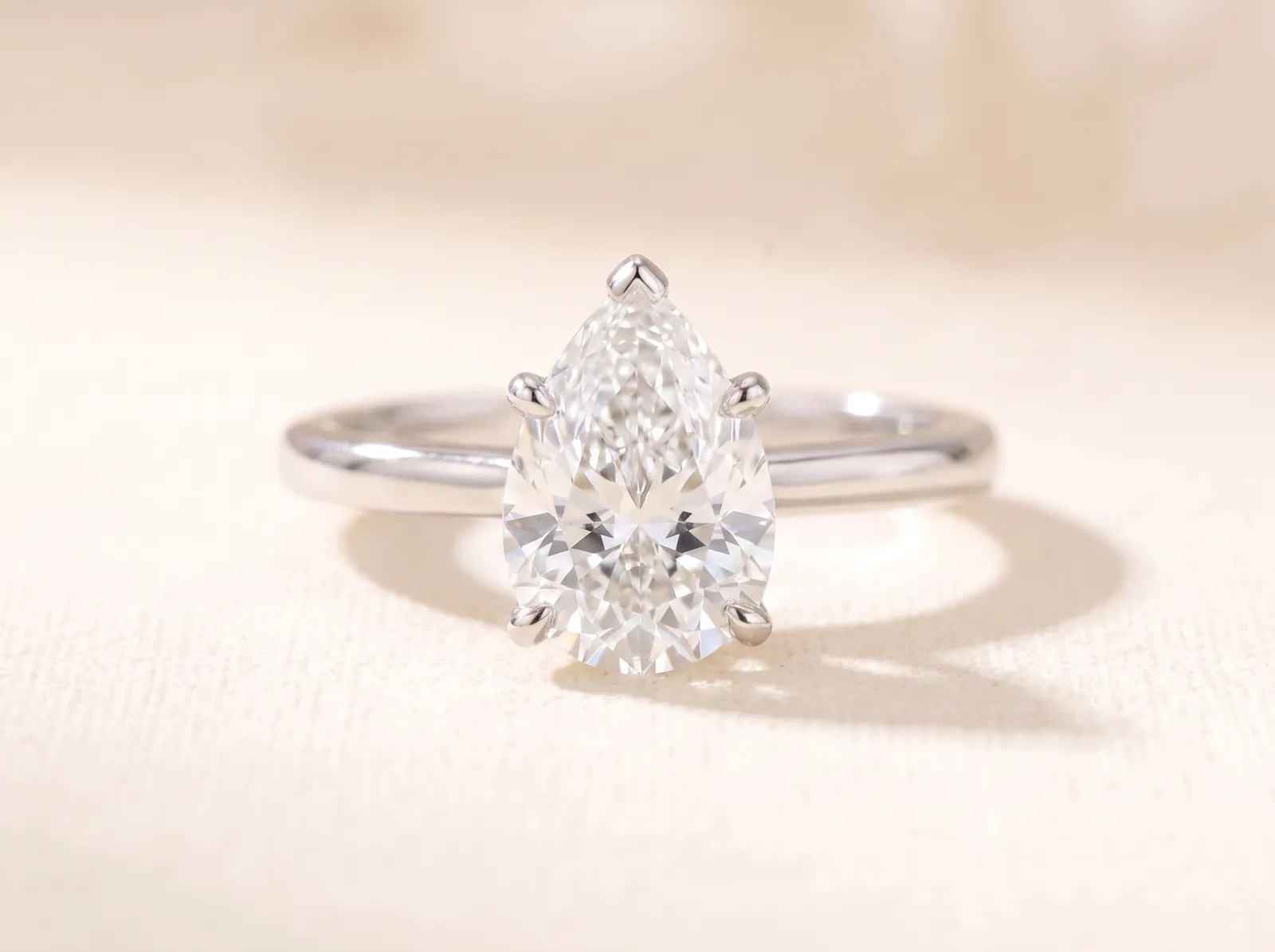 The same platinum solitaire lying on the cream surface, now holding a pear-shaped diamond, rounded at one end and drawn to a point at the other, facing the camera.