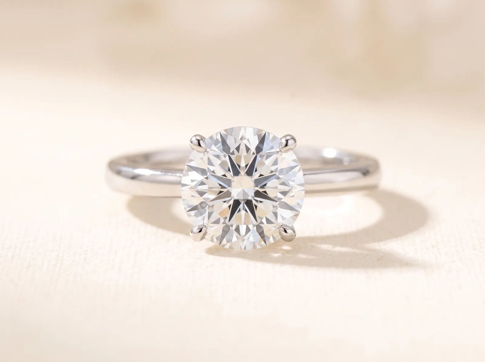 A platinum solitaire ring lying on a cream linen surface, a round brilliant diamond facing the camera in four slender prongs.