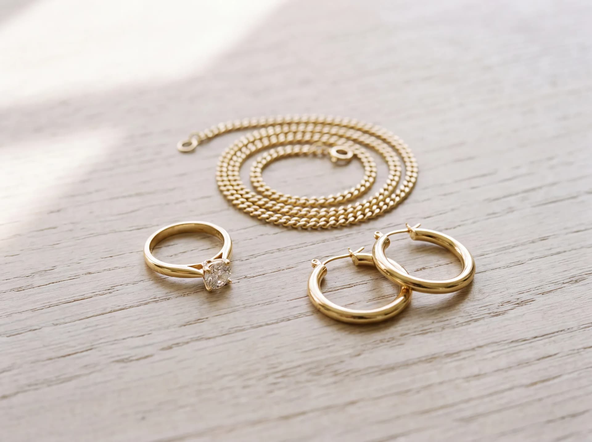 A finished gold ring, chain, and pair of hoop earrings arranged on a pale wood surface
