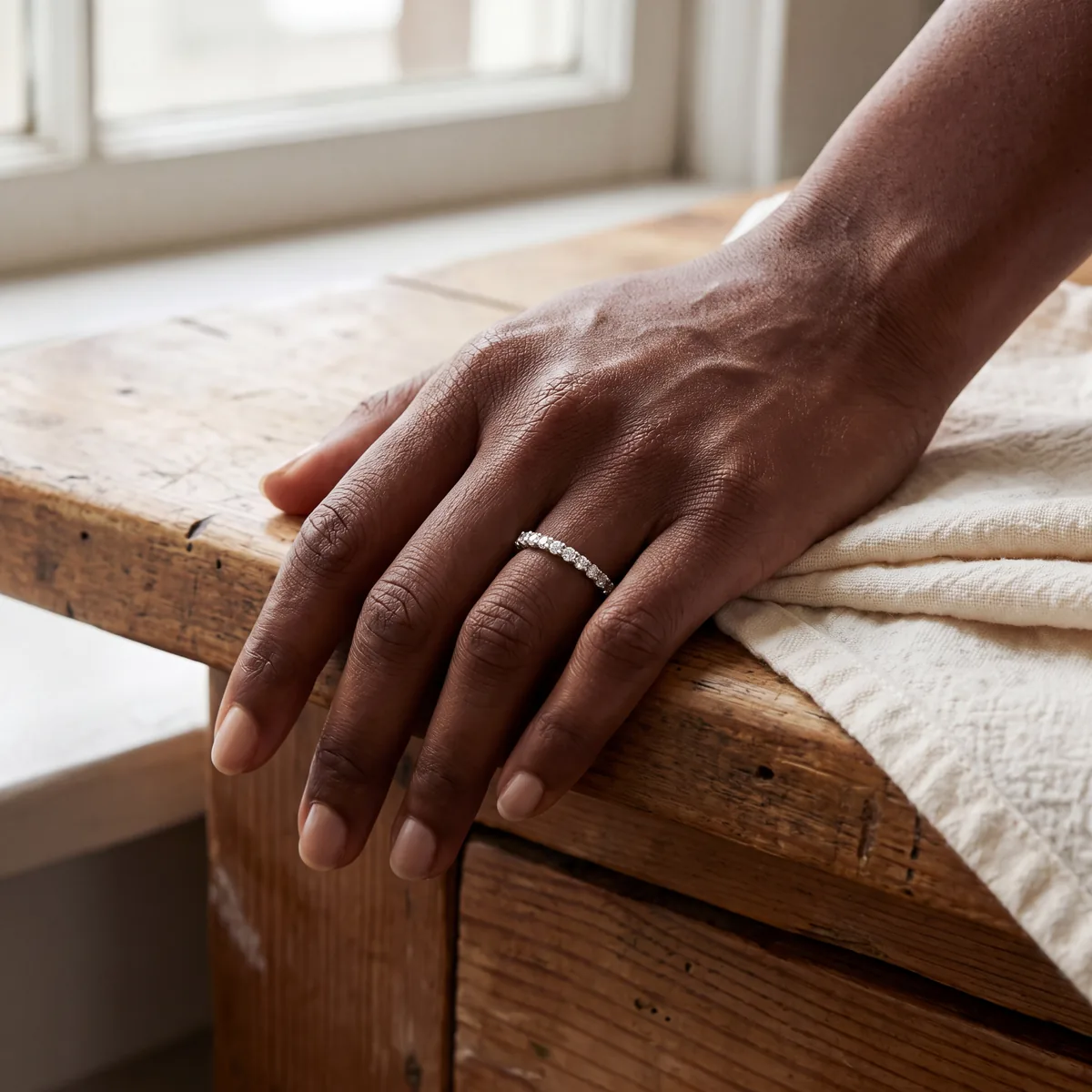 A hand resting on a worn wooden table beside a cream cloth, wearing a slim band set with a row of small diamonds.